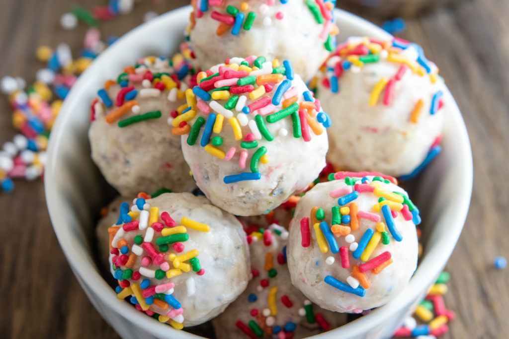 A photo of a bowl of birthday cake flavored protein balls. The balls are white with rainbow sprinkles on top. There are a few sprinkles on the side of the bowl. The background is a wooden surface.